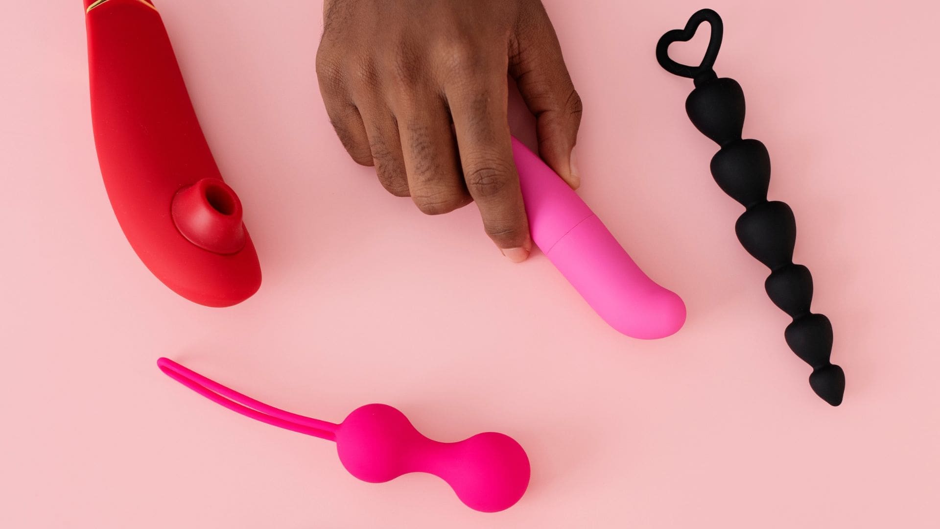 hand reaching out to hold beginner sex toys lying on a pink backdrop