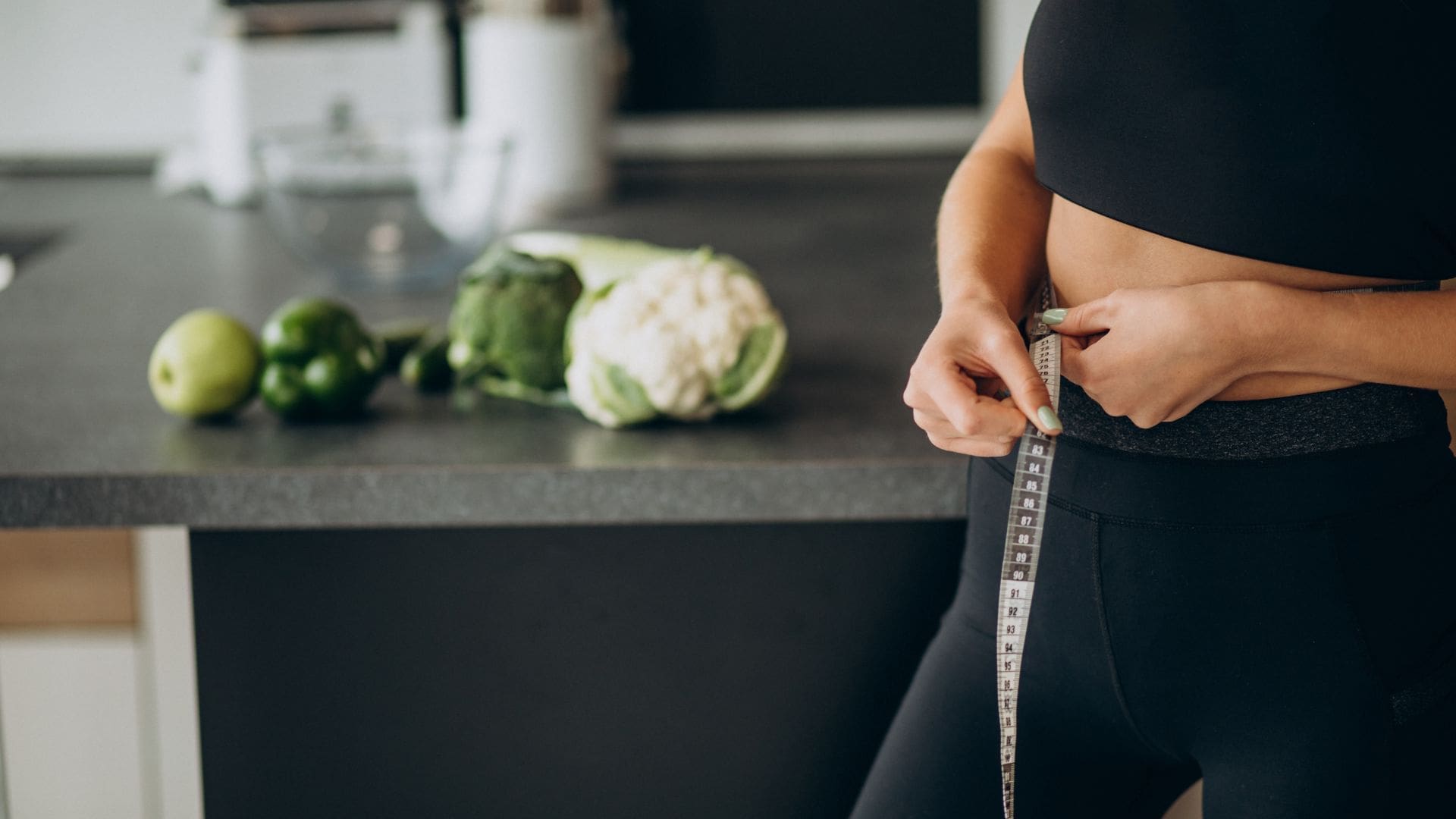 Woman wanting to know how to lose weight with a measuring tape around her waist in the kitchen with vegetables on the table