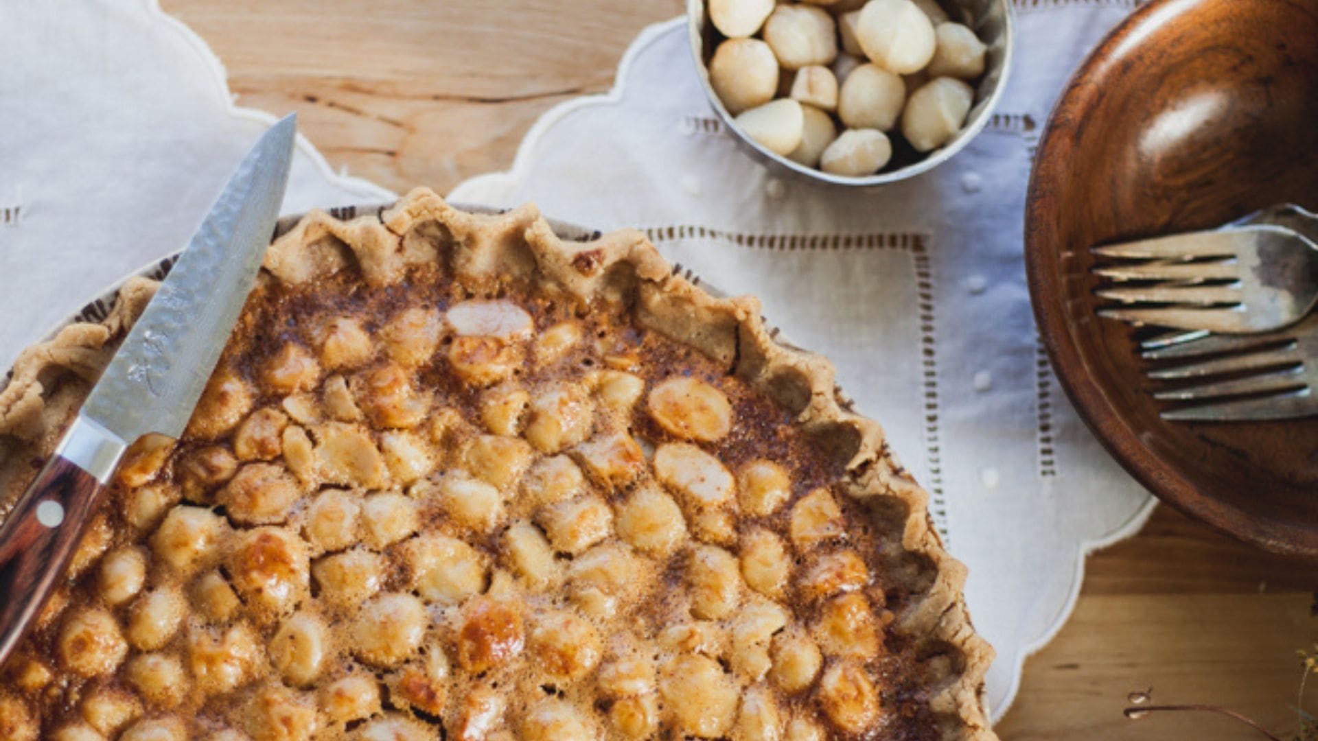 macadamia nut pie with knife balancing on top, a fork in a bowl and macadamia nuts in a bowl